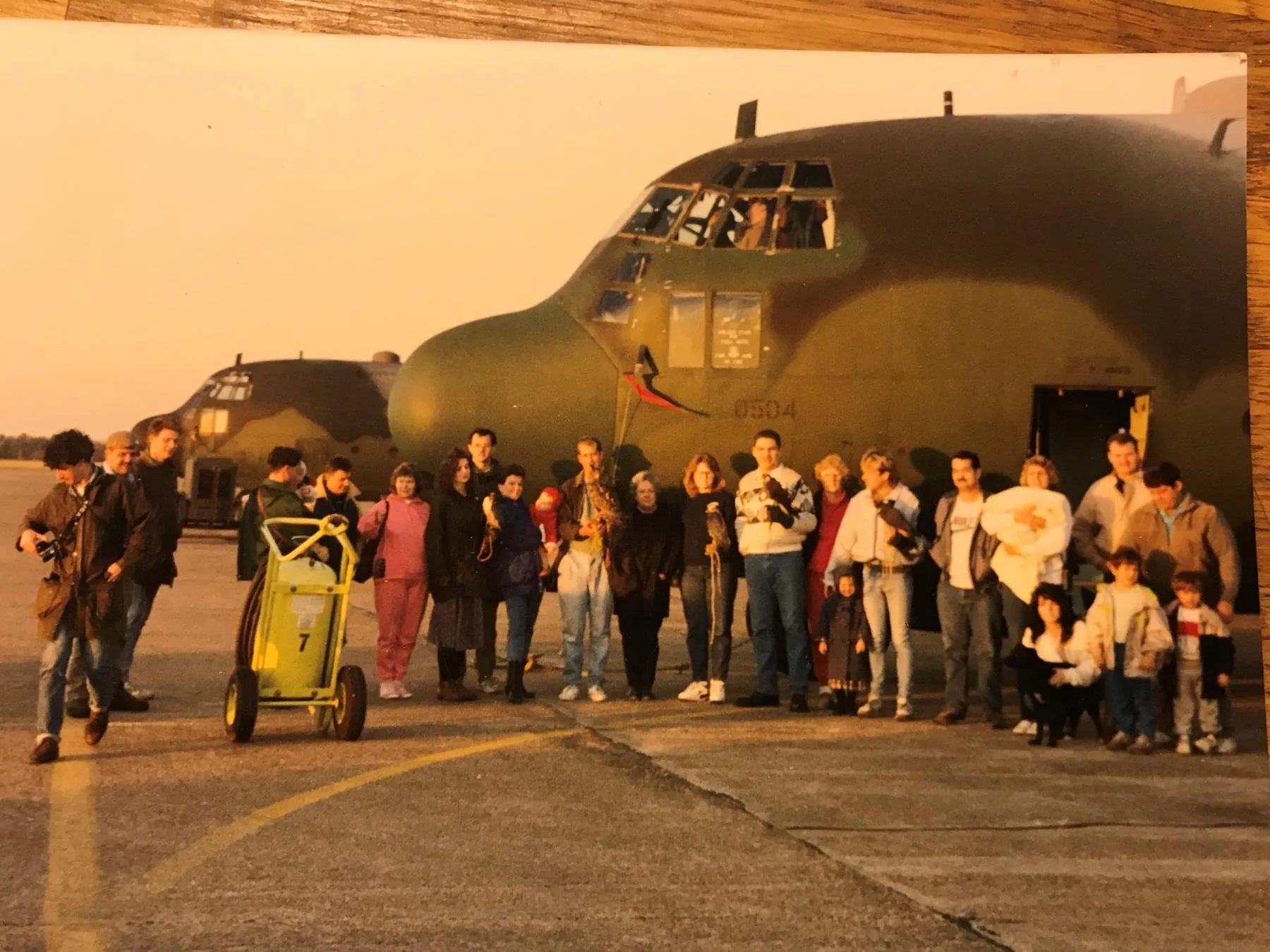 Group of Avian Environmental staff and handlers with falcons on the glove in front of a C-130 Hercules transport aircraft at golden hour — RAF airfield setting
